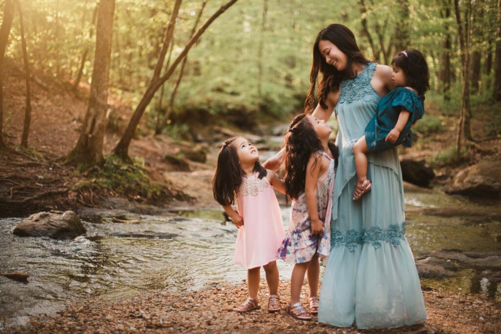 Mom standing with her three daughters beside the rocky creek bed surrounded by tall trees and woodland greenery.
