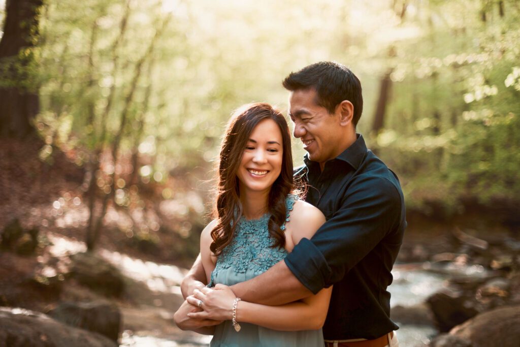Mom and dad embracing beside flowing creek water with warm sunlight filtering through the woods.