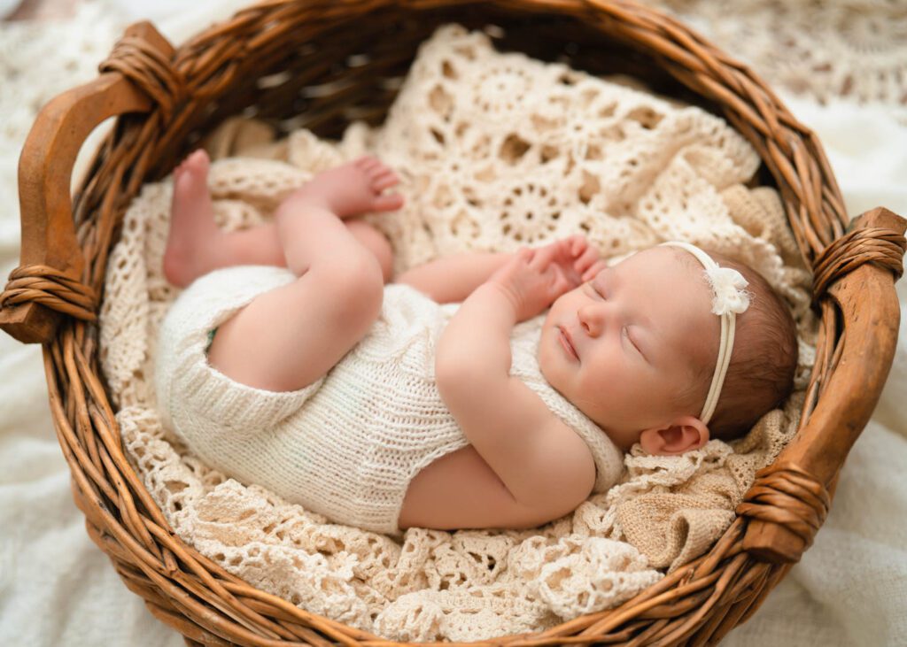Sleeping newborn baby girl in a wicker bowl on a vintage cream blanket wearing a white knit romper and bow headband.