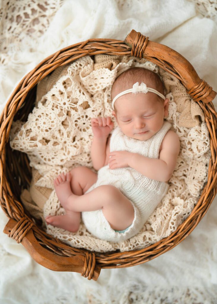 Sleeping newborn baby girl in a white knit romper and bow headband resting in a wicker bowl on crochet blankets.