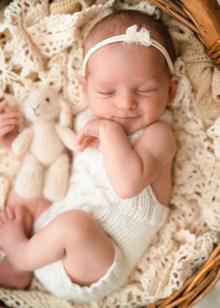 Smiling newborn baby girl holding a small bunny while wearing a cream knit romper on a vintage crochet blanket.