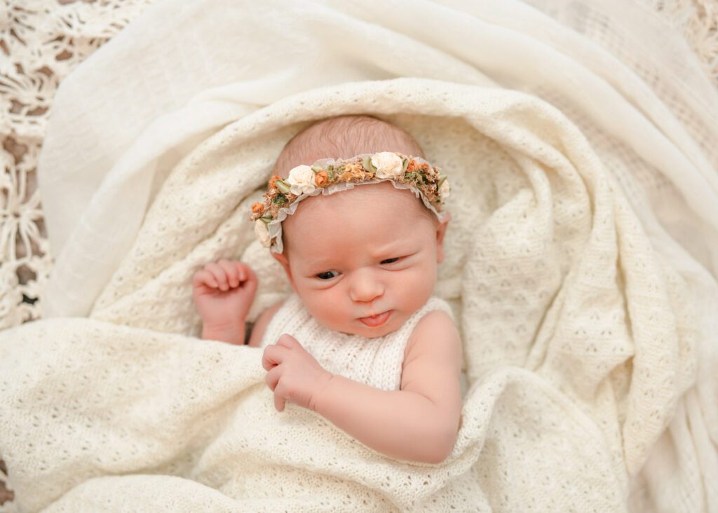 Newborn baby girl making a playful expression on a cream blanket while wearing a floral crown