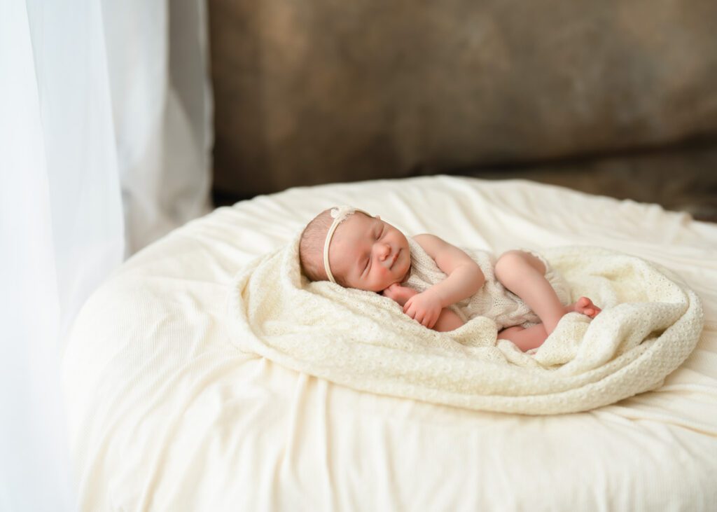 Newborn baby girl lying on a vintage blanket by a window in a cream knit outfit and bow headband.