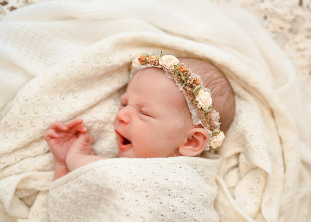 Newborn baby girl yawning on a cream knit blanket while wearing a soft floral headband.