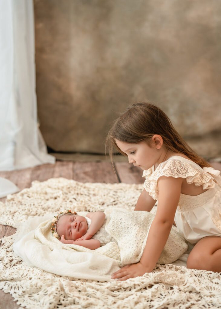 Big sister looking down at her newborn sister as both wear cream outfits beside a bright window.