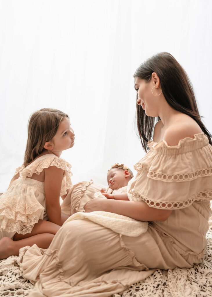 Mom sitting on the floor holding her newborn daughter as big sister looks up at her beside a bright window.