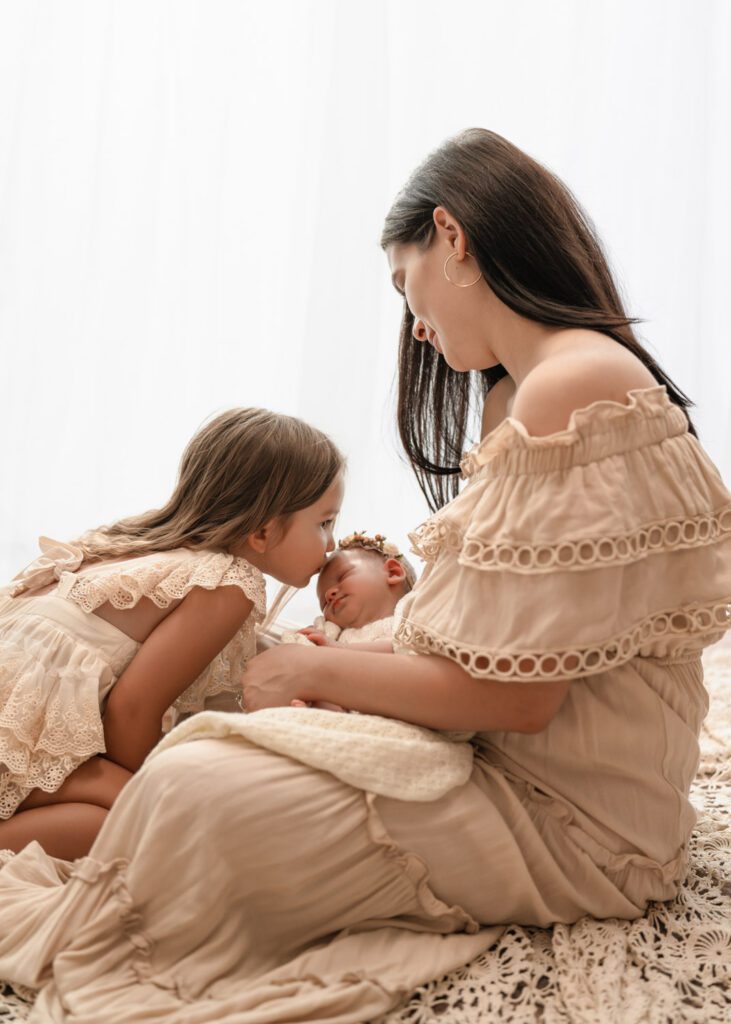 Mom sitting with her two daughters beside a bright window as big sister kisses the newborn on the head.