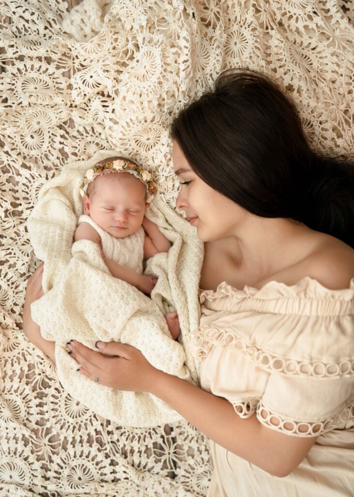 Mother looking at her newborn daughter as they rest together on a vintage crochet blanket in cream outfits.