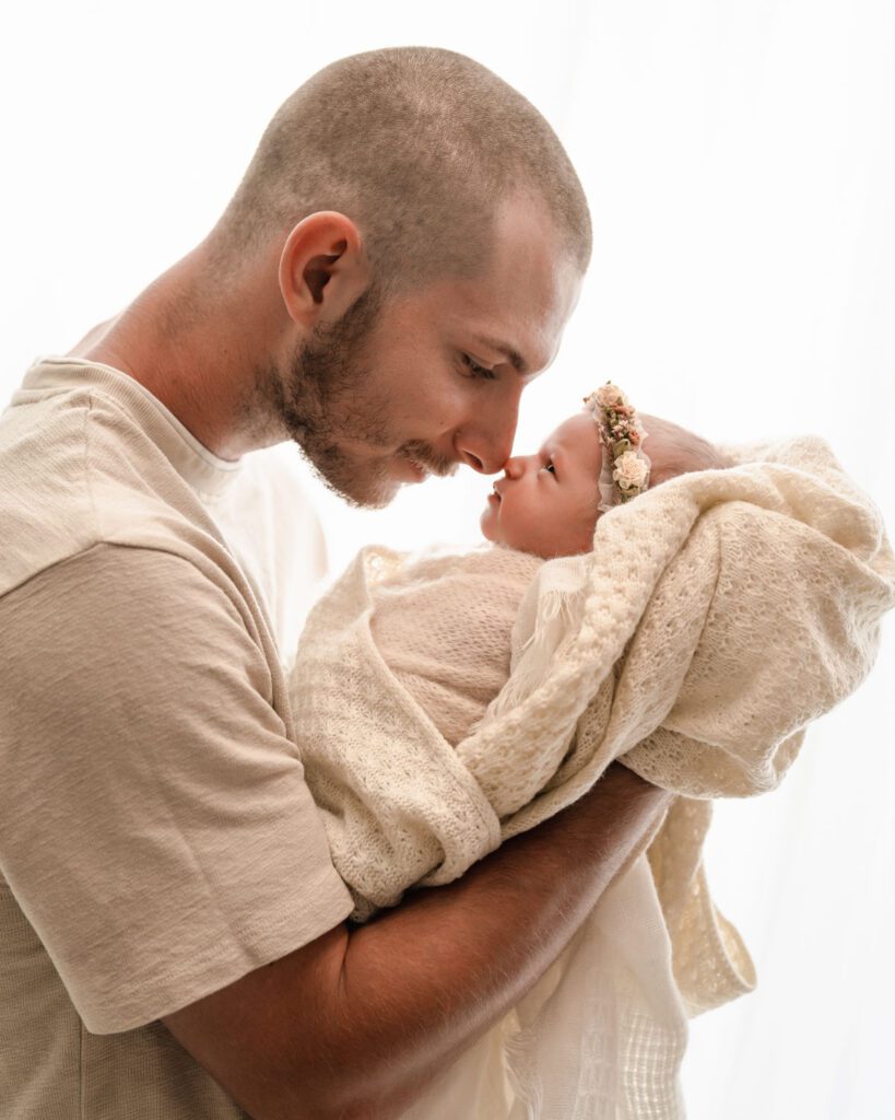 Father holding his newborn daughter nose to nose as they look toward each other beside a bright window.