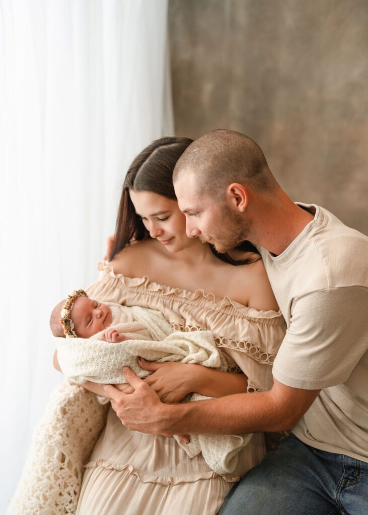 Mom and dad sitting together in beige while looking down at their newborn daughter for a Roswell newborn photographer.