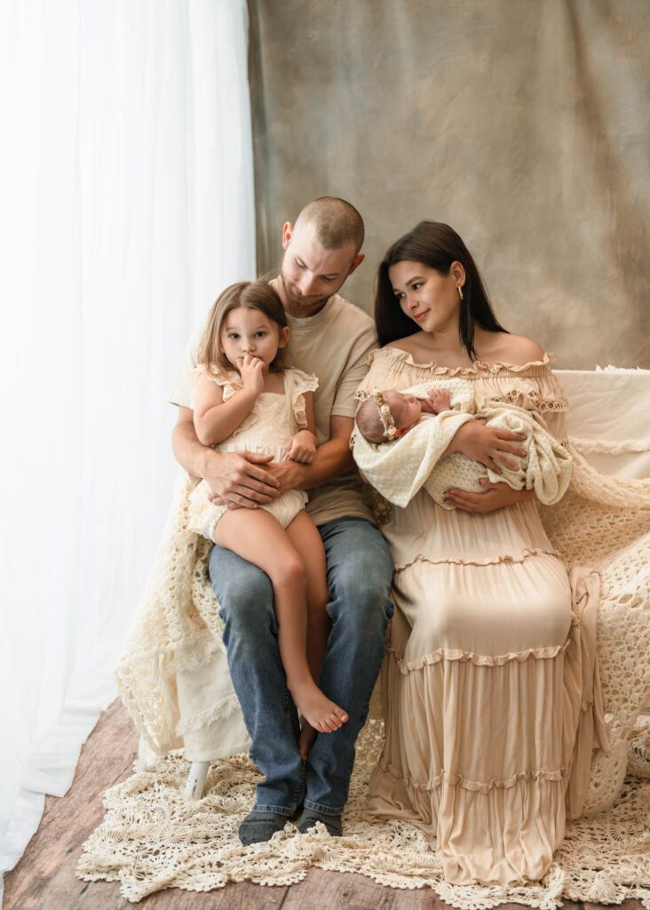 Family cuddling beside a bright window as mom holds the newborn and dad holds big sister for Roswell newborn photos.