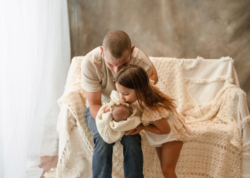 Dad sitting beside a window with his newborn daughter as big sister kisses the baby on the head.