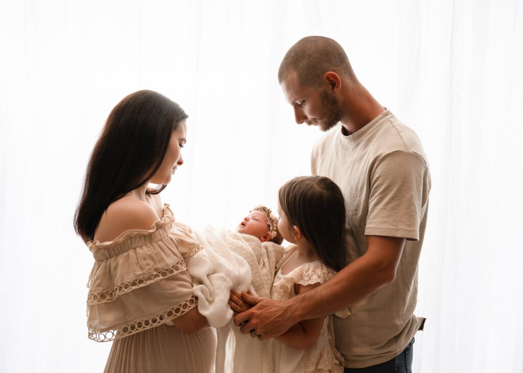 Family standing by a bright window looking down at their newborn baby girl during a studio newborn session.