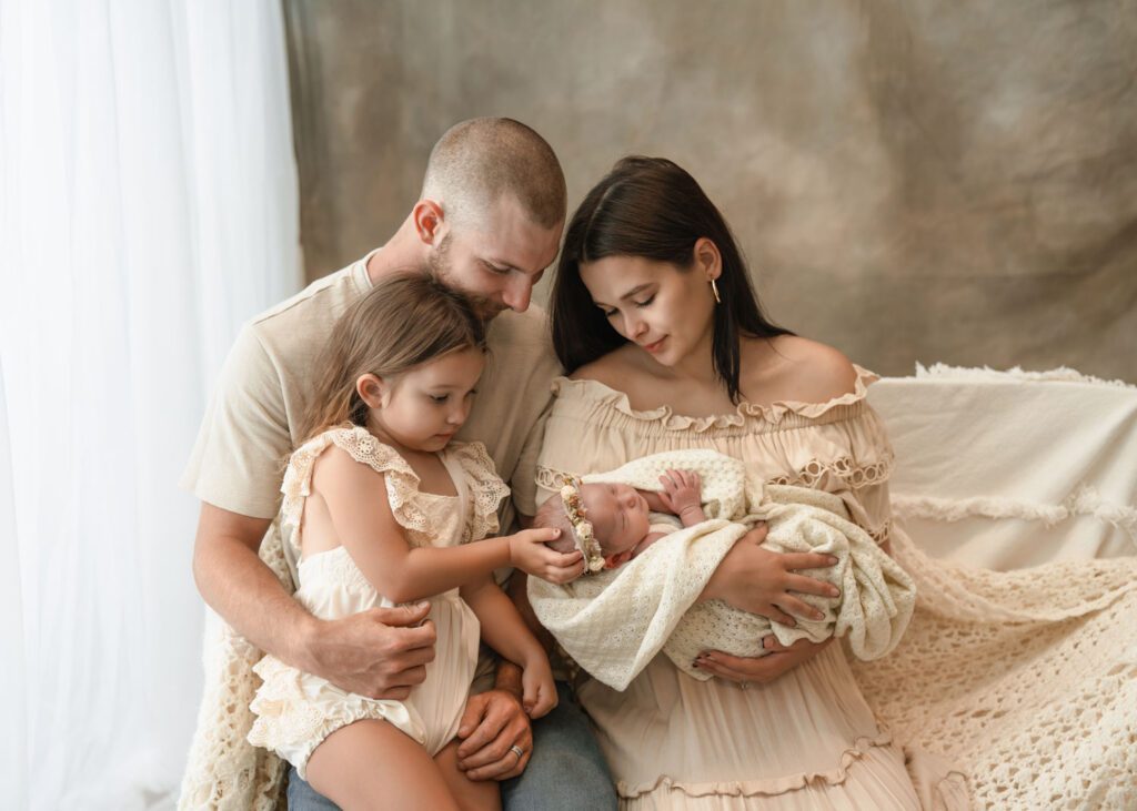 Big sister gently touching her newborn sister’s head while mom and dad gather close beside them.