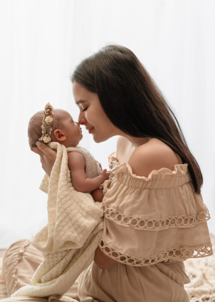 Mom holding her newborn daughter nose-to-nose in a beige eyelet dress as baby wears a floral headband.