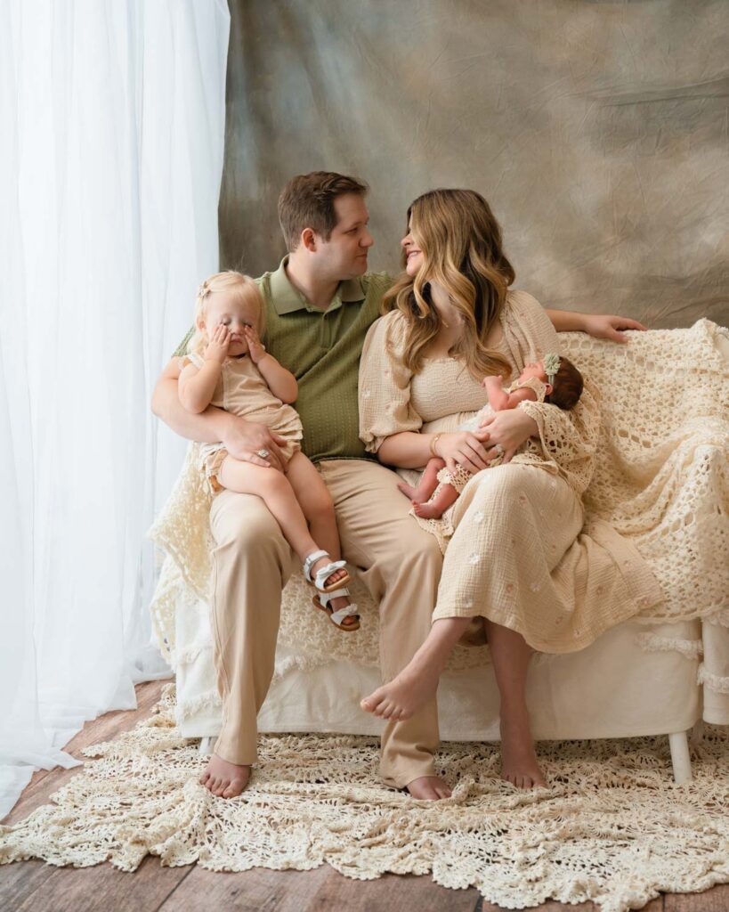 Mom and dad looking at each other while holding their newborn baby and sleepy toddler daughter.