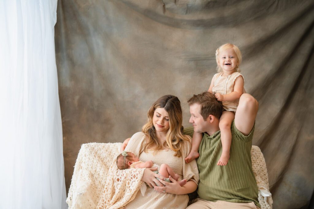 Family of four cuddling together as mom holds their newborn baby and big sister smiles from dad’s shoulders.