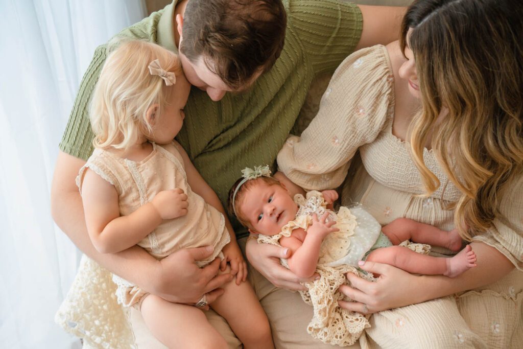 Family sitting together as dad holds their toddler daughter and mom cradles their curious newborn baby.