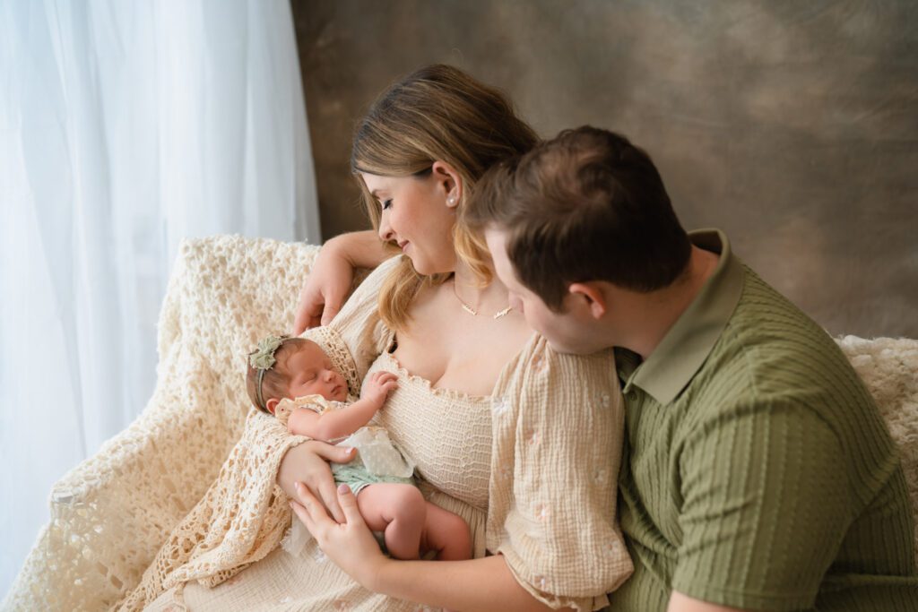 Parents looking down at their newborn baby girl while seated together beside a bright window.