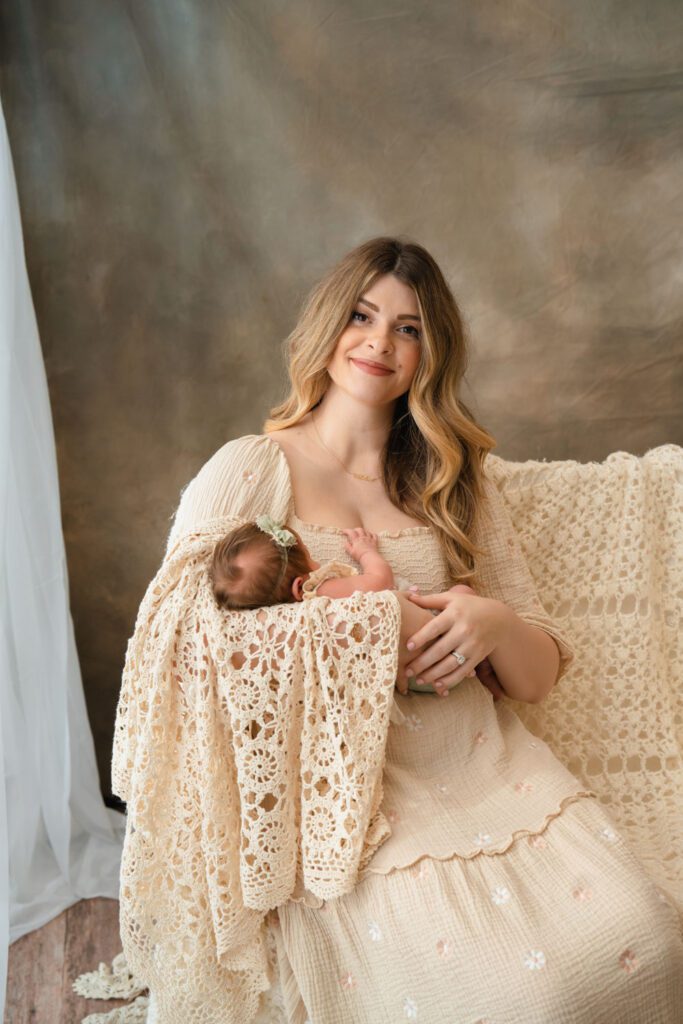 Mom in a beige floral dress holding her newborn daughter wrapped in crochet beside a bright window.