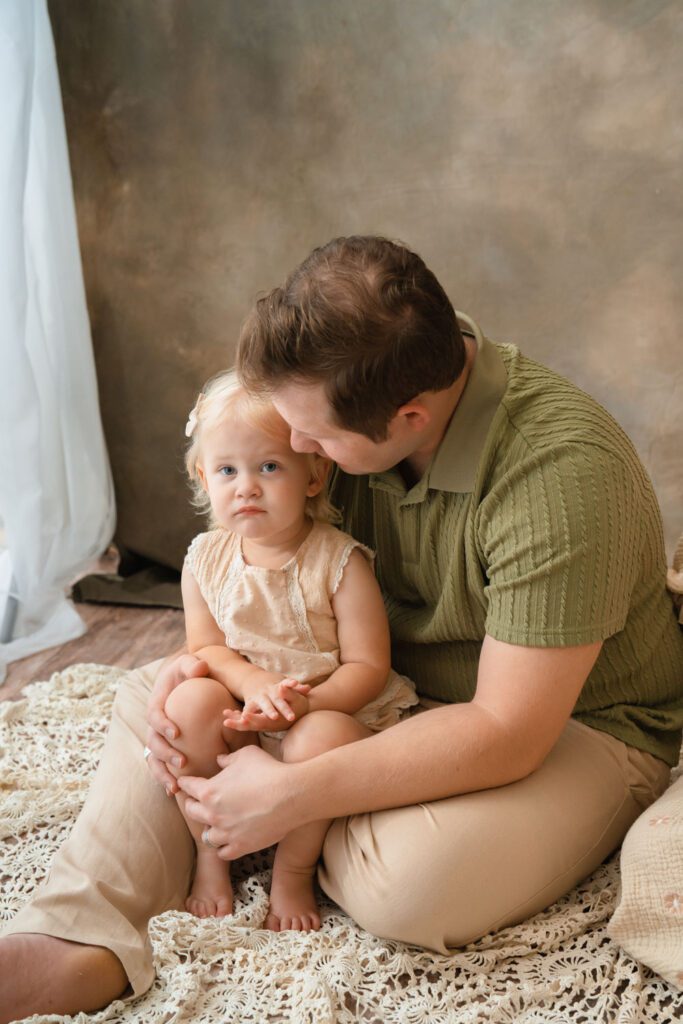 Dad holding his blonde toddler daughter in his lap while sitting on the floor against a warm brown backdrop.