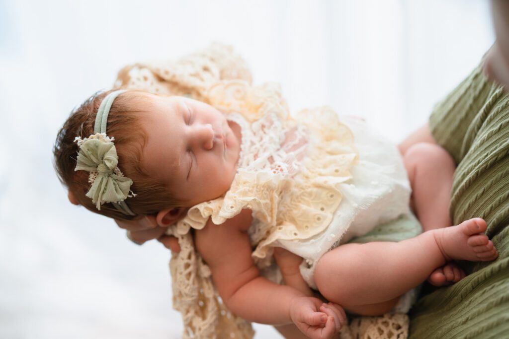 Sleeping newborn baby girl in a cream lace romper cradled in dad’s hands beside a bright window.