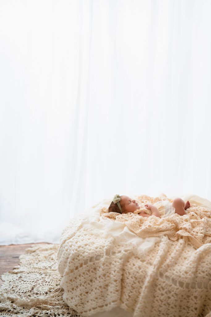 Newborn baby girl lying on layered cream crochet blankets in a softly lit room with white curtains.