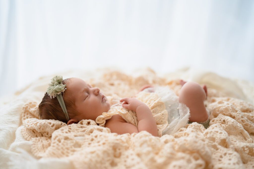 Newborn baby girl in a cream and green lace outfit resting on a vintage blanket with one hand on her chest.