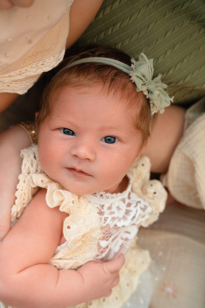 Alert newborn baby girl in lace outdit looking toward the camera while resting in her mom’s arms.