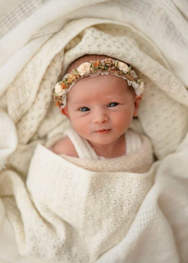 six day old Awake newborn baby laying on white crochet blankets and wearing a floral headband.