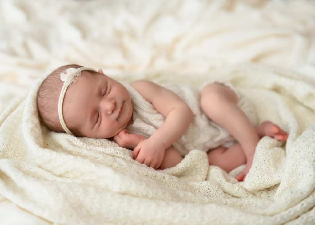 six day old Newborn baby girl wearing a white crochet romper with headband laying on knit cream blankets. 