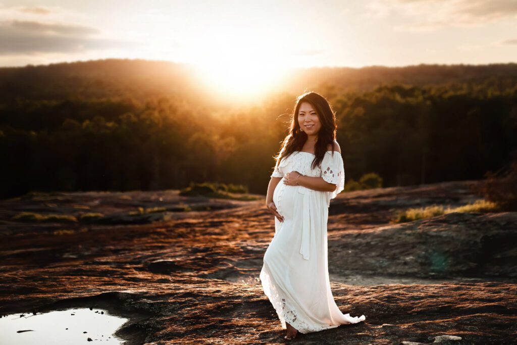 Pregnant woman in a white gown standing alone on monadnock with a glowing Arabia Mountain sunset behind her.