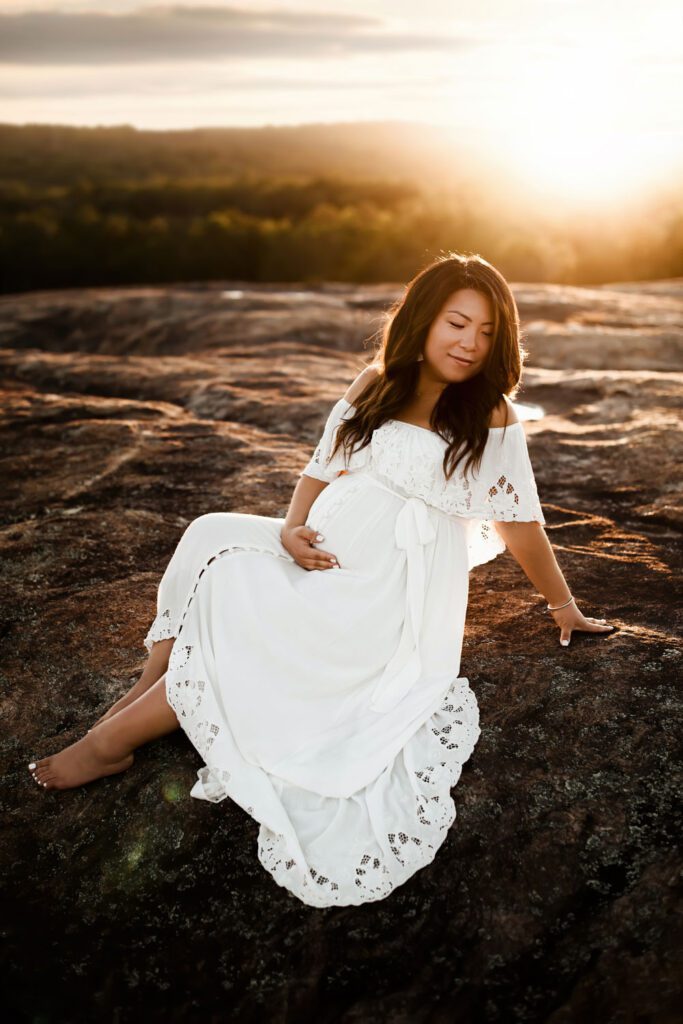 pregnant mother reclining on big rocky area in the mountains in a white eyelet dress at golden hour.