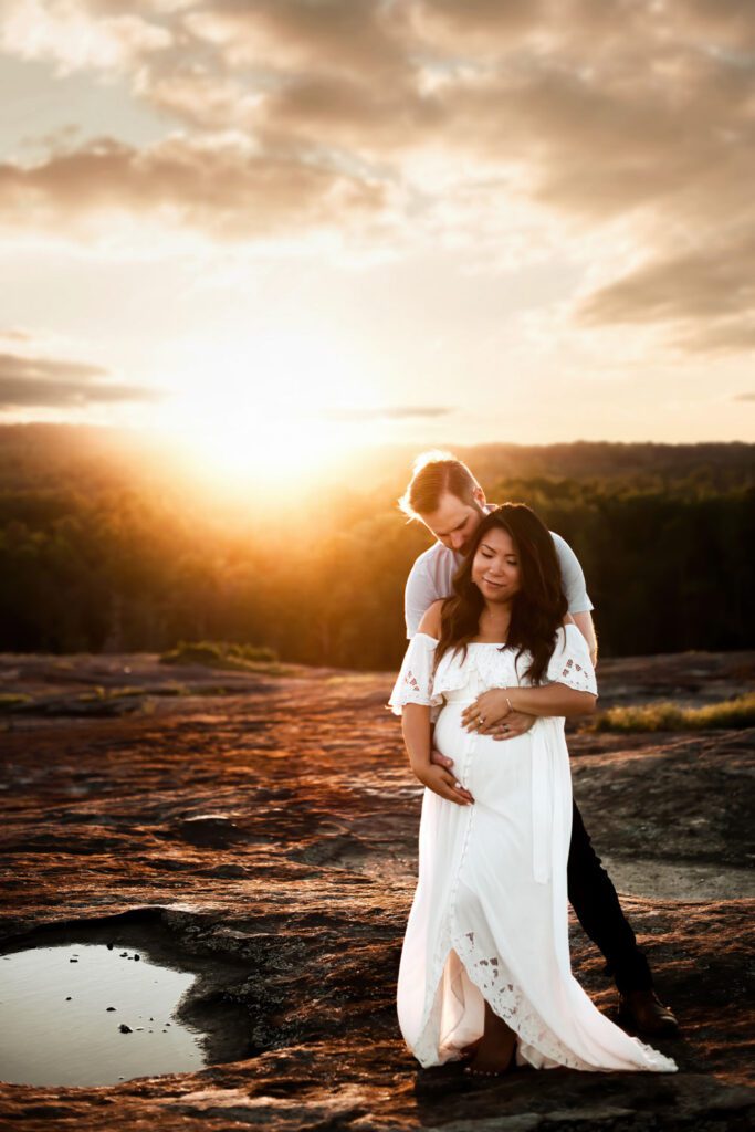pregnant mom in a white gown cradling her belly while husband stands behind her in warm Stonecrest sunset light.