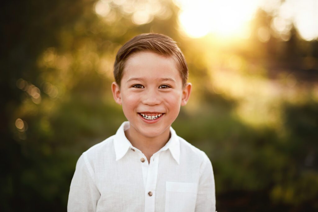 Smiling boy in a white shirt photographed in warm golden light with softly blurred greenery behind him.