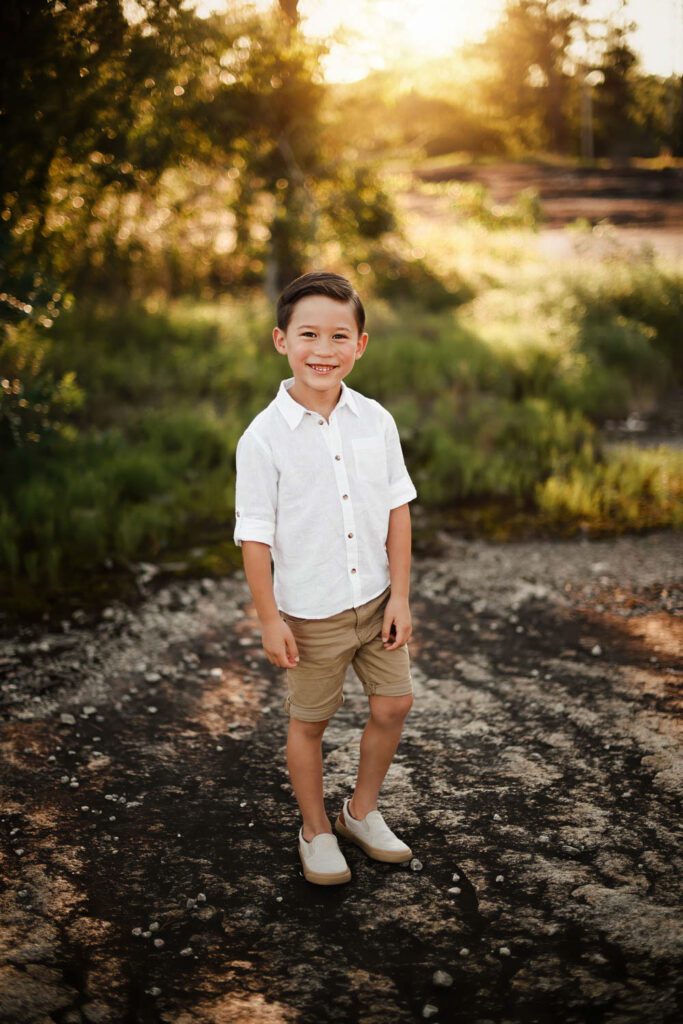 Smiling boy standing on rocky terrain with sunlit greenery in the background during an outdoor portrait session.