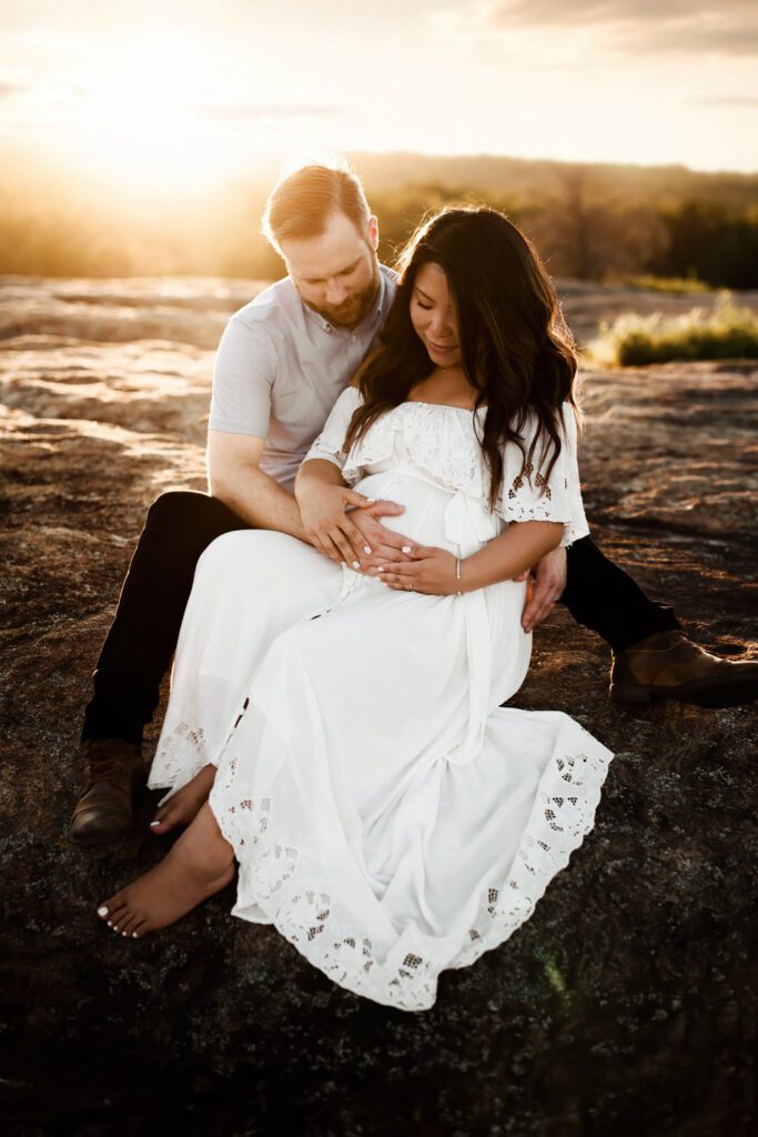 Pregnant woman in a white gown seated beside husband with both hands resting around her belly on the rocky ground.