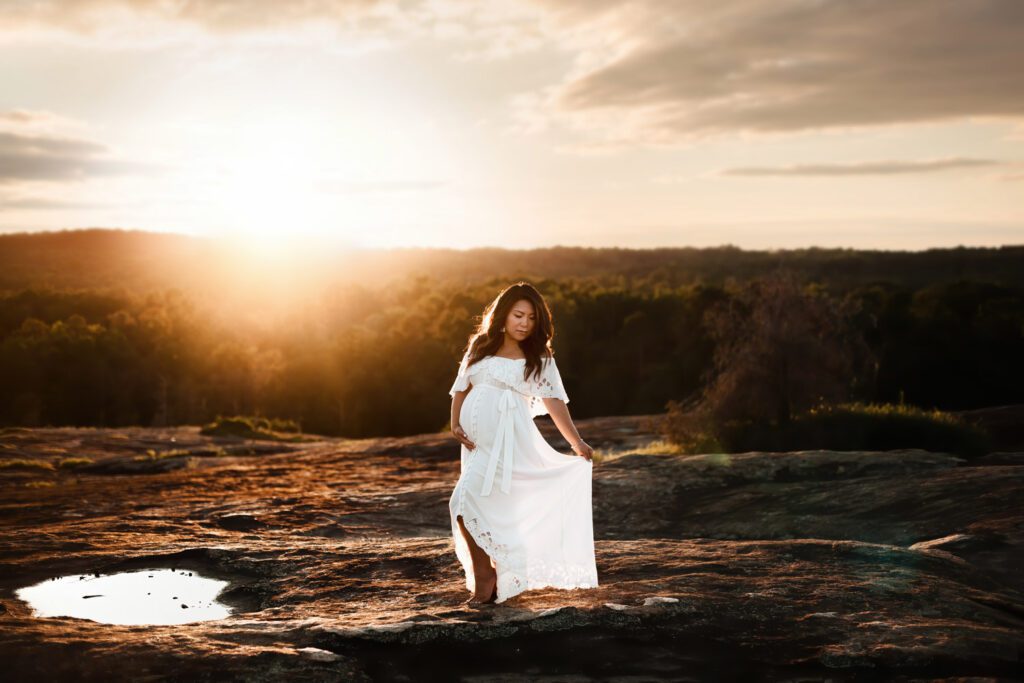 Pregnant woman in white gown standing alone at sunset with the  Arabia Mountain Mile Rock Boardwalk in the background in Stonecrest, GA.
