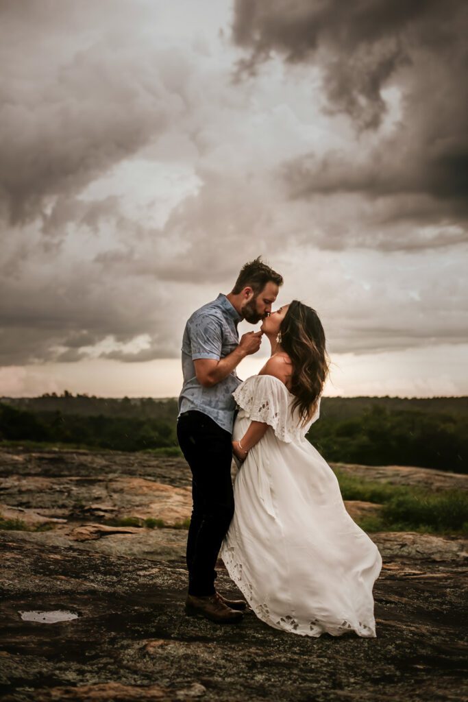 Pregnant woman and her partner kissing on wet rocks beneath stormy clouds as he gently touches her chin.