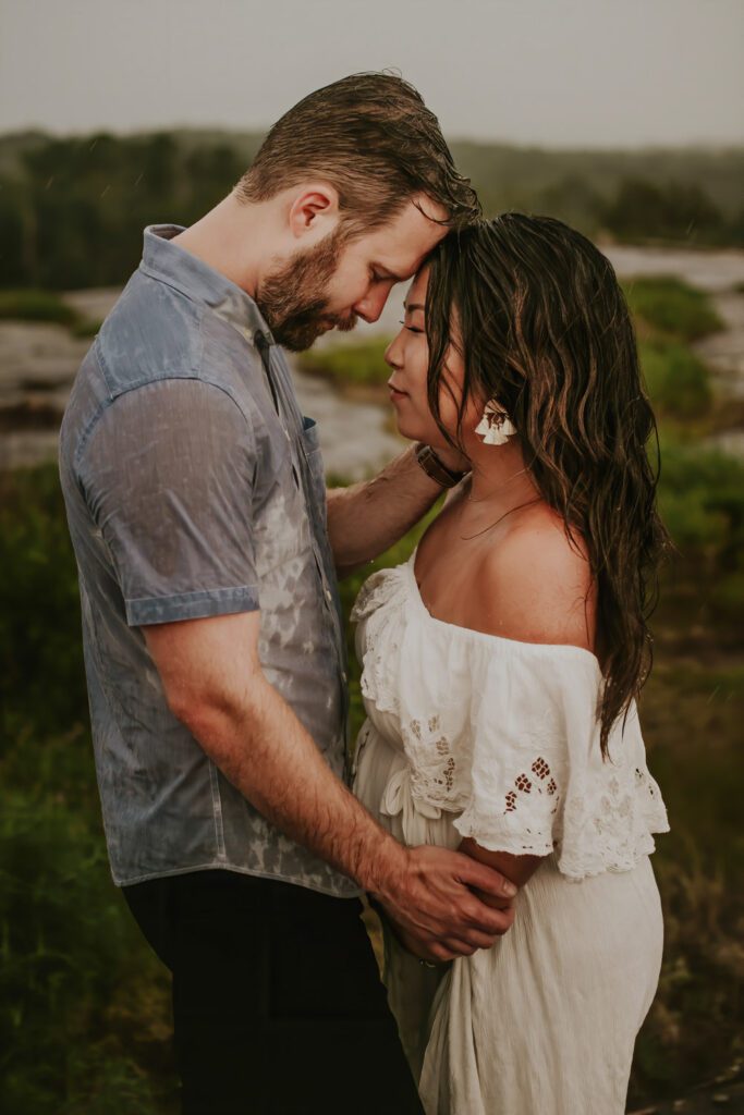 expecting couple facing each other wet from the rain touching foreheads during maternity photos outdoors.