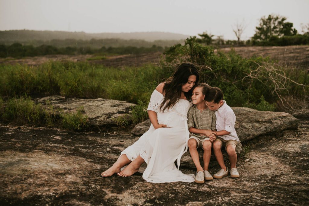 Pregnant woman in a white gown seated on the Diamorpha Pools with two sons beside her under a cloudy stormy sky.
