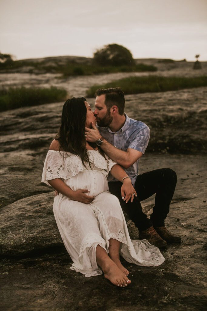 Expecting mother in a white gown seated on Diamorpha Pools as her partner leans close beside her during a rainy session.