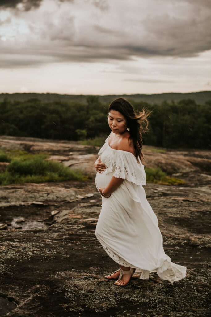 Pregnant woman in a white off-the-shoulder gown cradling her belly on rocky terrain beneath a cloudy sky.