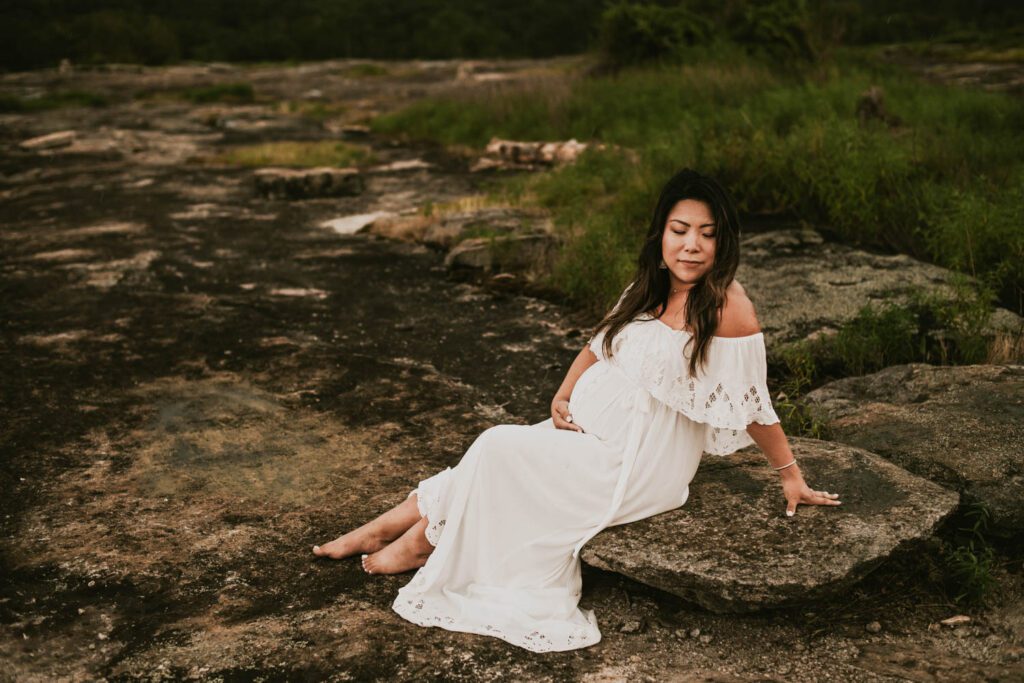 Expecting mother in a white gown sitting beside the rock cairns at Arabia Mountain in Stonecrest, GA