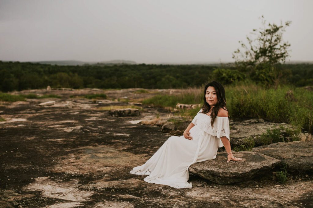 Pregnant woman in a flowing white gown seated on wet rocks at Arabia Mountain with her dress draped around her.