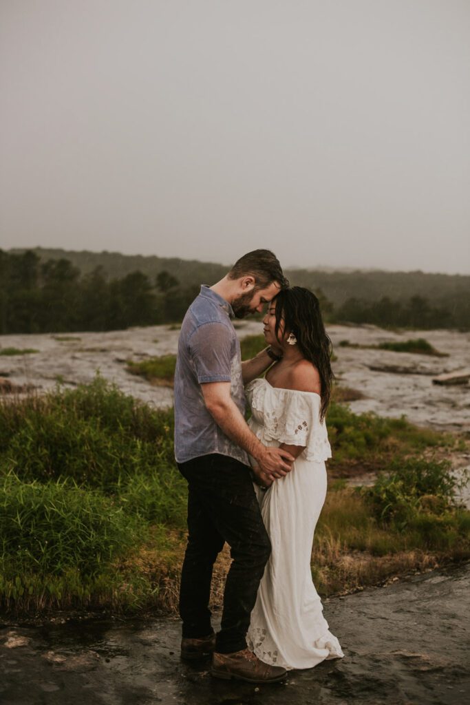 Expecting mother in a white gown standing close with her partner on wet rocky terrain under an overcast sky.