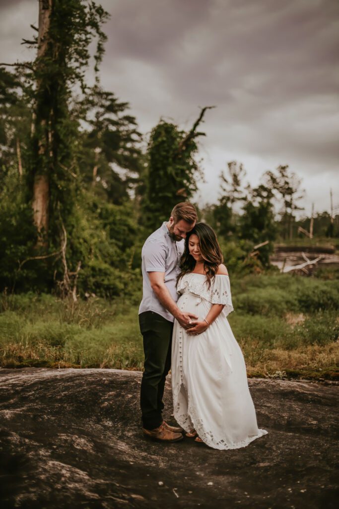 Pregnant woman in a white gown standing with her husband on swirling tidal gray rocks at Arabia Mountain in Stonecrest.
