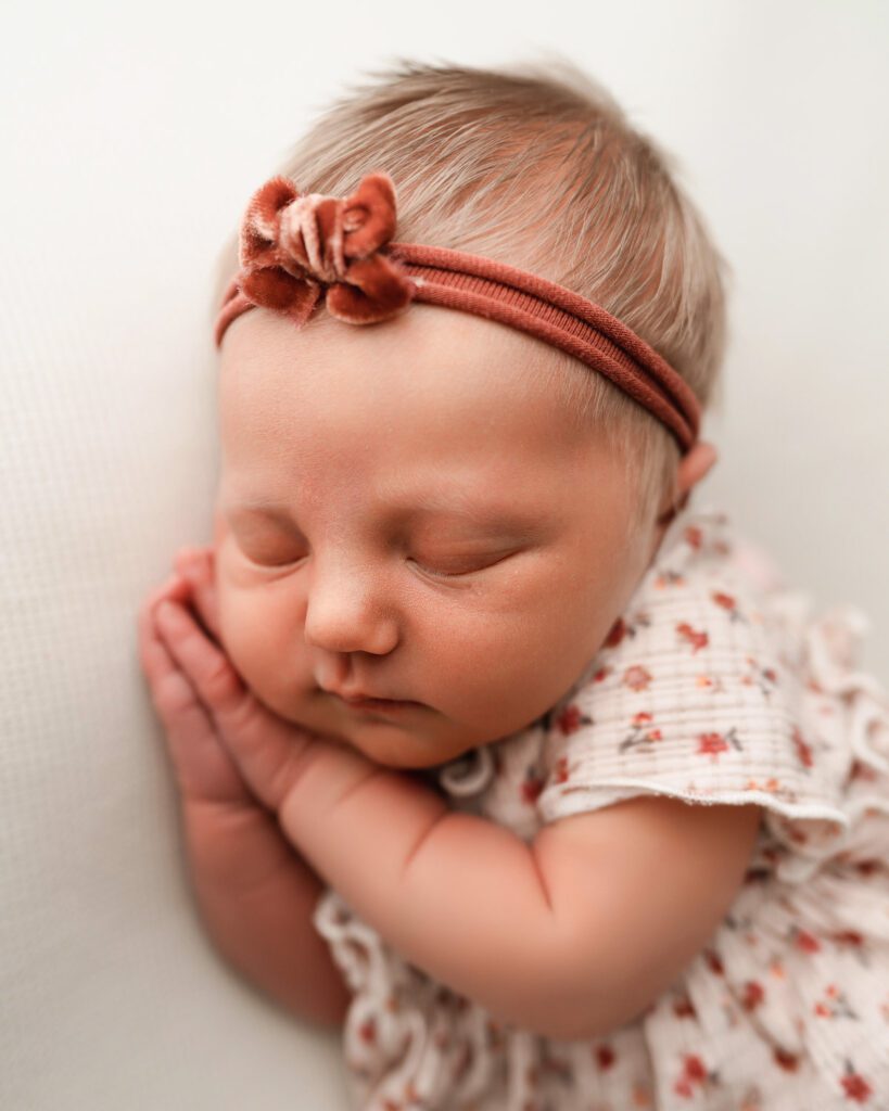 three week old baby girl with hands under her cheek wearing a pink floral outfit with a bow headband in her blonde hair. 