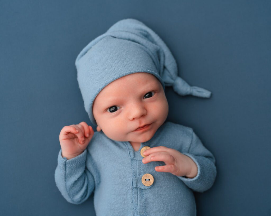 two week old baby boy wearing blue sleeper onesie and matching hat laying on a blue fabric background. 
