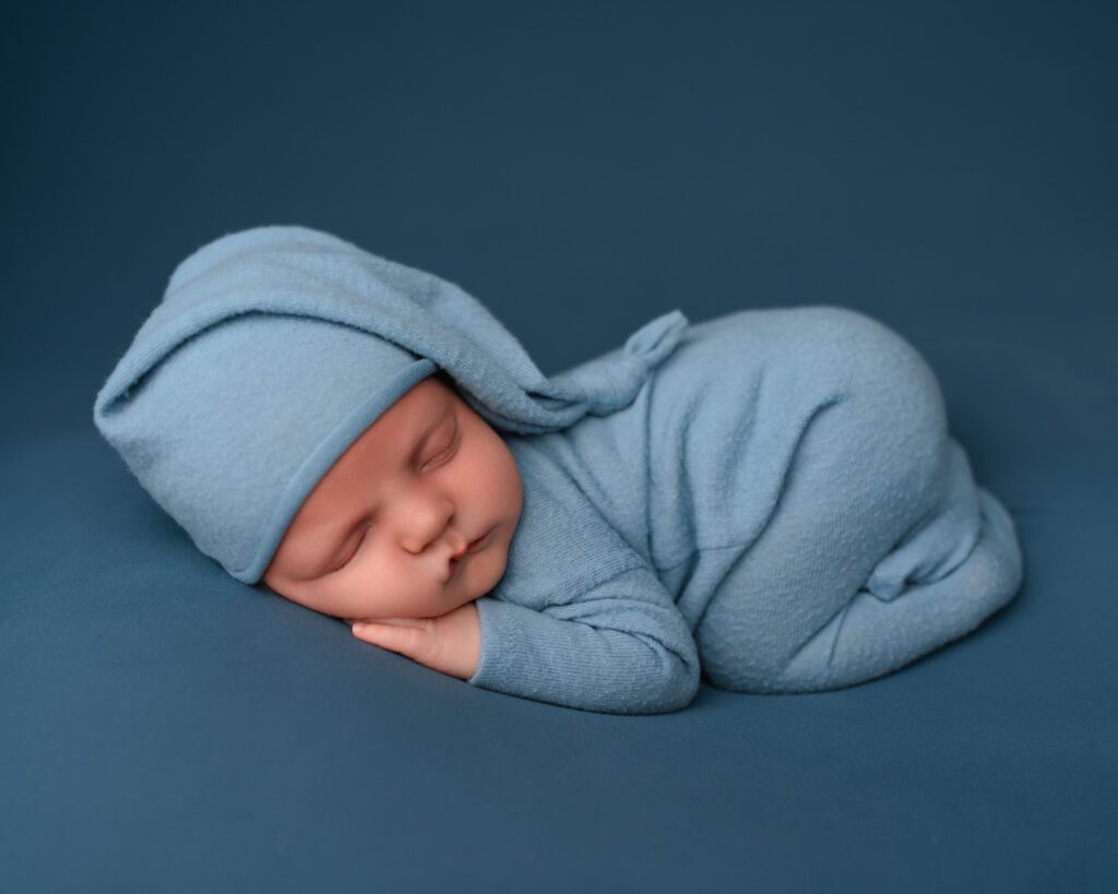 Baby boy dressed in blue fabric onesie with a blue knotted sleepy cap hat while laying on his stomach on a blue backdrop. 